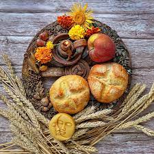 An arrangement of two baked buns, stalks of wheat, an apple, a candle, and small yellow, red, and orange flowers