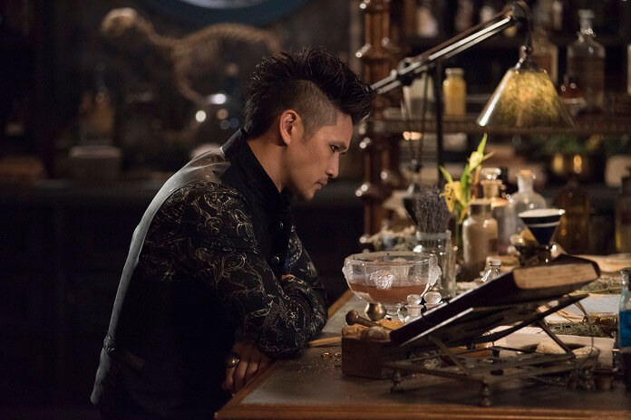 Magnus Bane dressed in black sitting at a table covered in magical items from the TV show Shadowhunters.