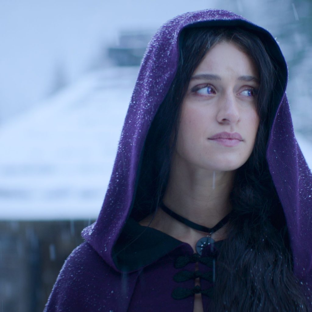 Yennefer of Vengerberg from the TV show "The Witcher" wears a purple robe and looks off to the side with a snowy cabin in the background. 