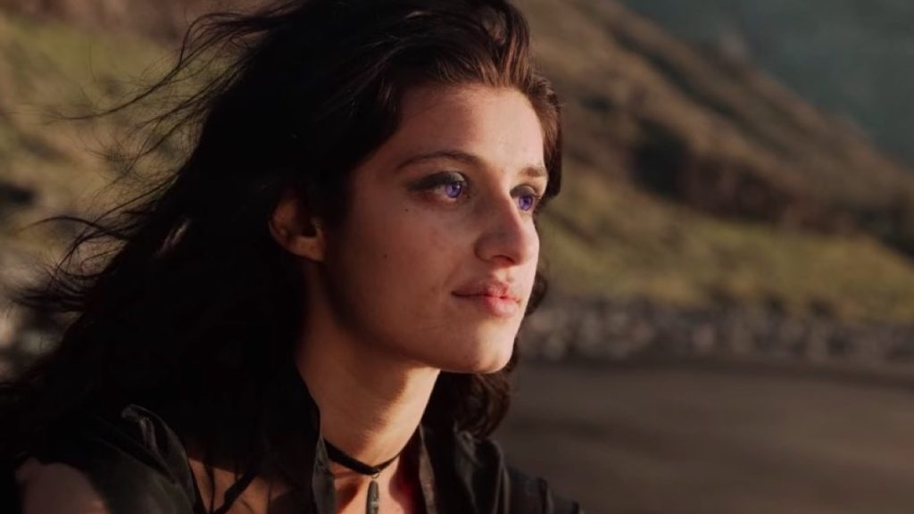 Yennefer of Vengerberg from the TV show "The Witcher" sits on a beach and looks out at the waves with a dejected countenance. 