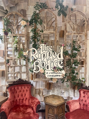 Wall of books with wood window frames in front, a sign that says "the ripped bodice a romance bookstore" in front/ Two velvet red chairs and table sit below it