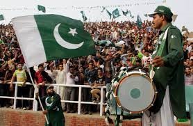 Parade for Pakistan Independence Day and one man is holding the Pakistan flag while another is playing a drum with a large crowd in the background
