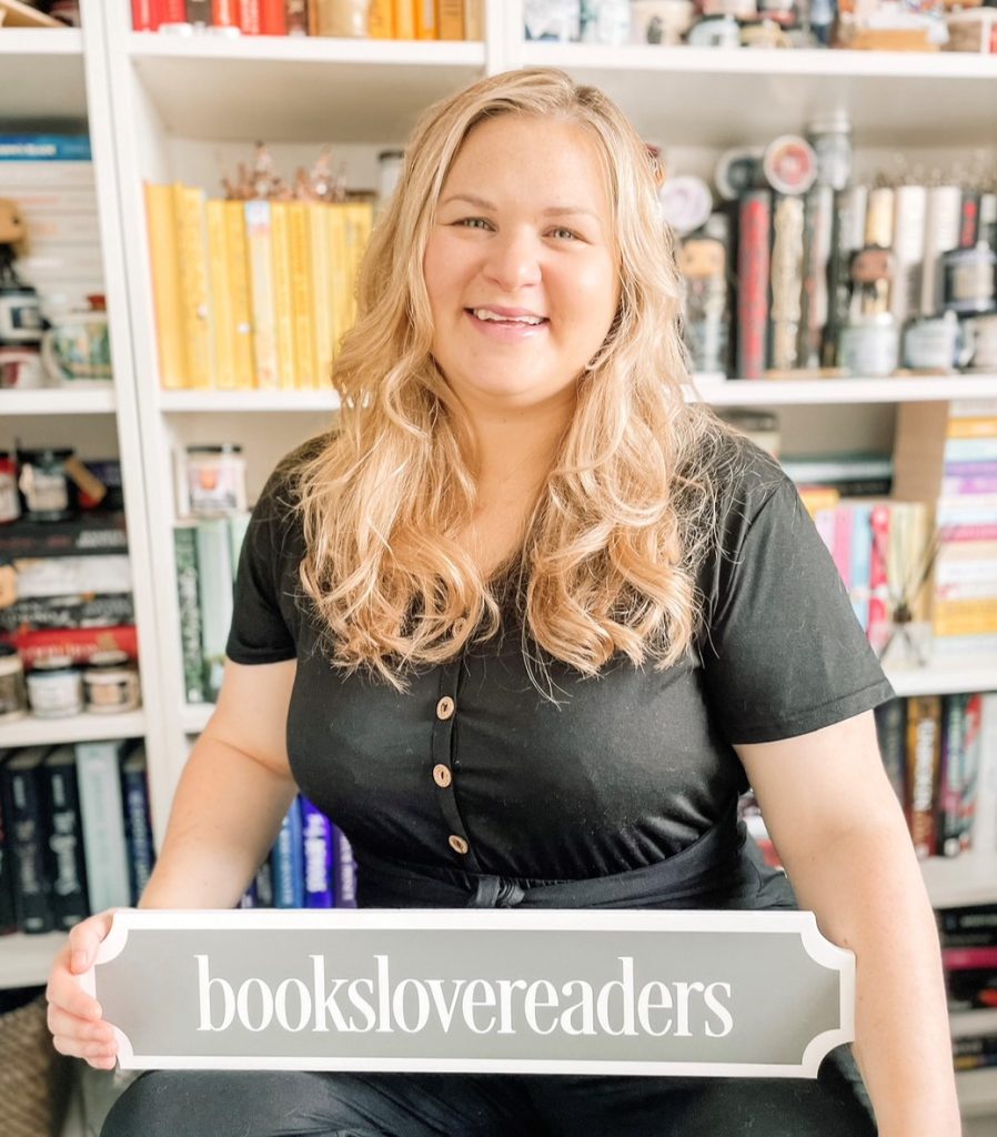 Stephanie smiling in front of a colorful bookshelf. She's holding a sign that reads "Bookslovereaders"