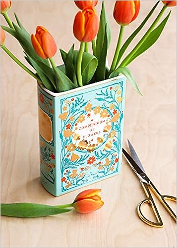 A blue ceramic book vase with orange tulips is in it. The book vase is has orange and red flowers with golden foil on them and the middle of the vase says a compendium of flowers