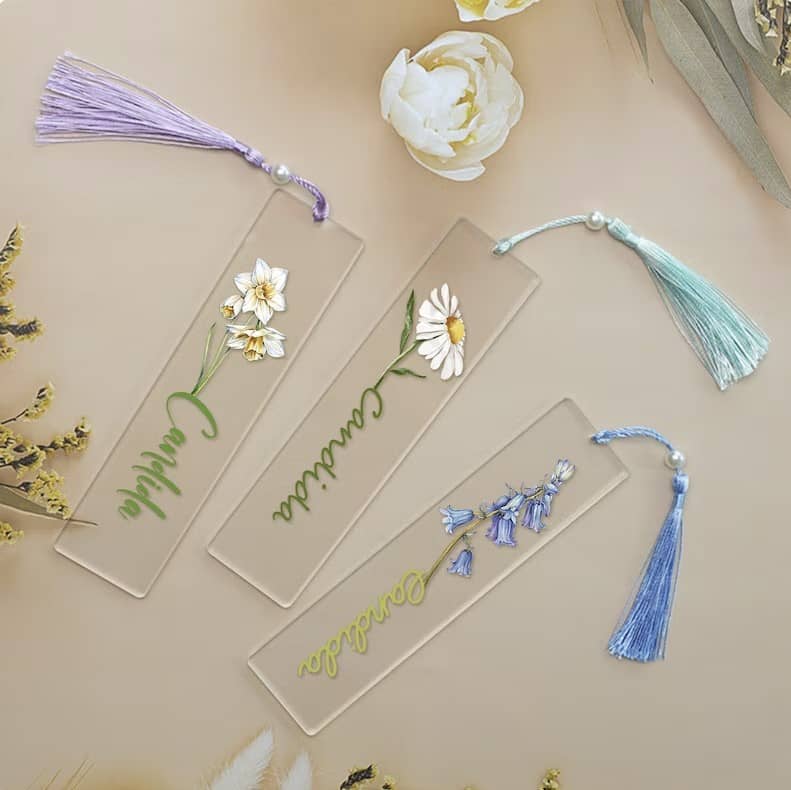 Three bookmarks with different colored tassels sit on a table. On the bookmarks are names in different fonts and colors connected to different flowers.