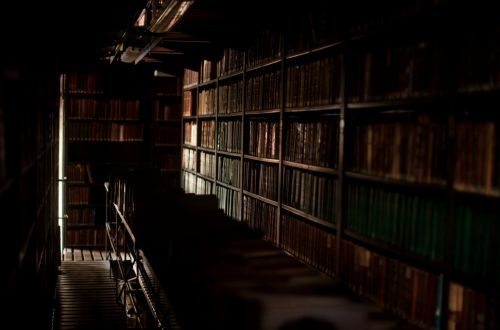 A dark library with very old books and chairs with a long table to read. 