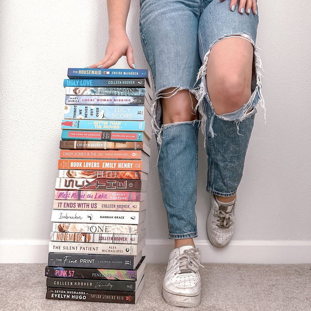Bookstagrammer Sara McCoy stands donning blue ripped denim and white sneakers, next to a knee-high pile of books.