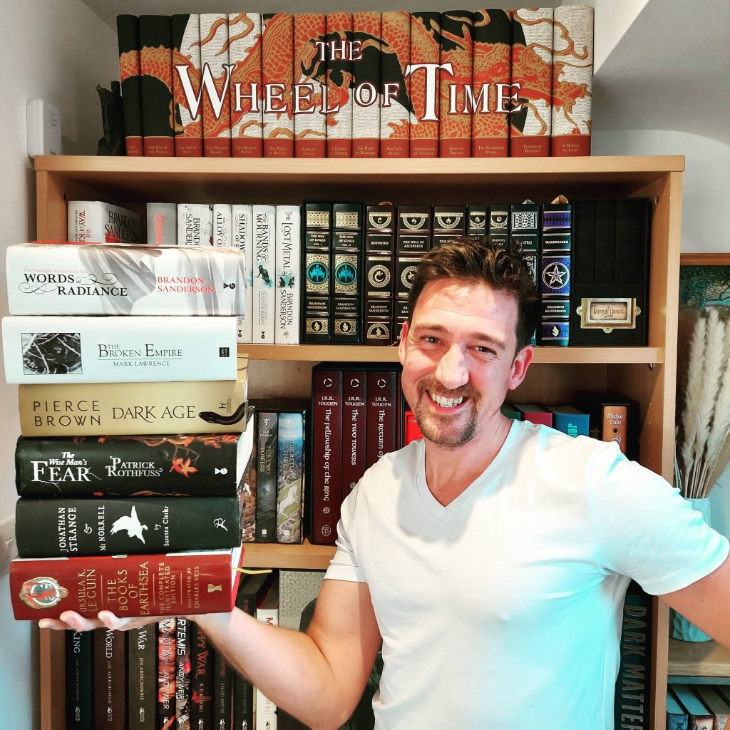 James of @fantasyphile holding up a stack of fantasy books in front of an organized bookshelf