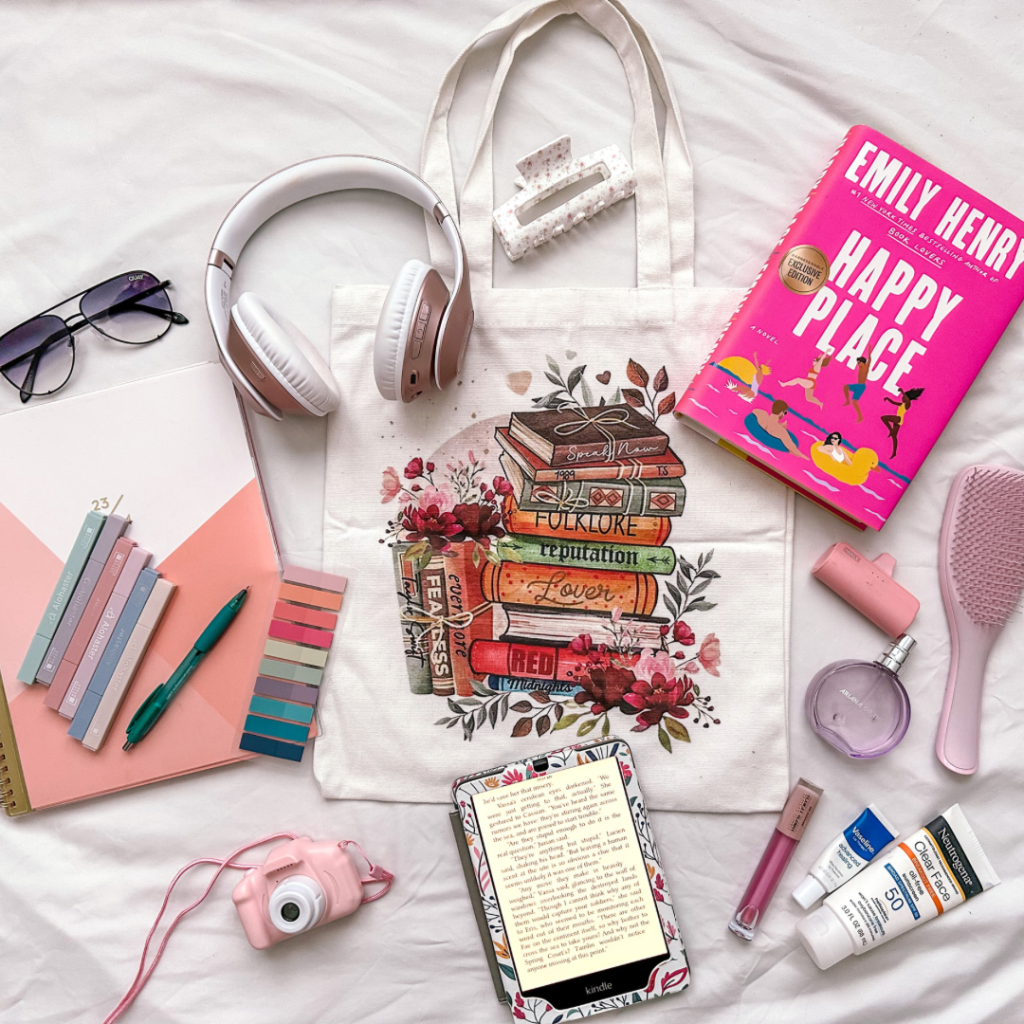 A beautiful flatlay of white headphones, shaded aviators, a pink and white camera, Emily Henry's book Happy Place, a bookish tote, a white clutcher, and e-book reader, and some more interesting paraphernalia, put together by Bookstagrammer Sara.
