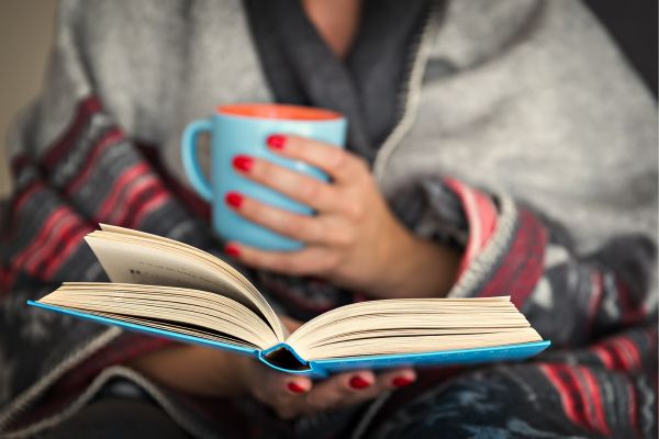 Woman reading a book cozied up in a blanket and mug.