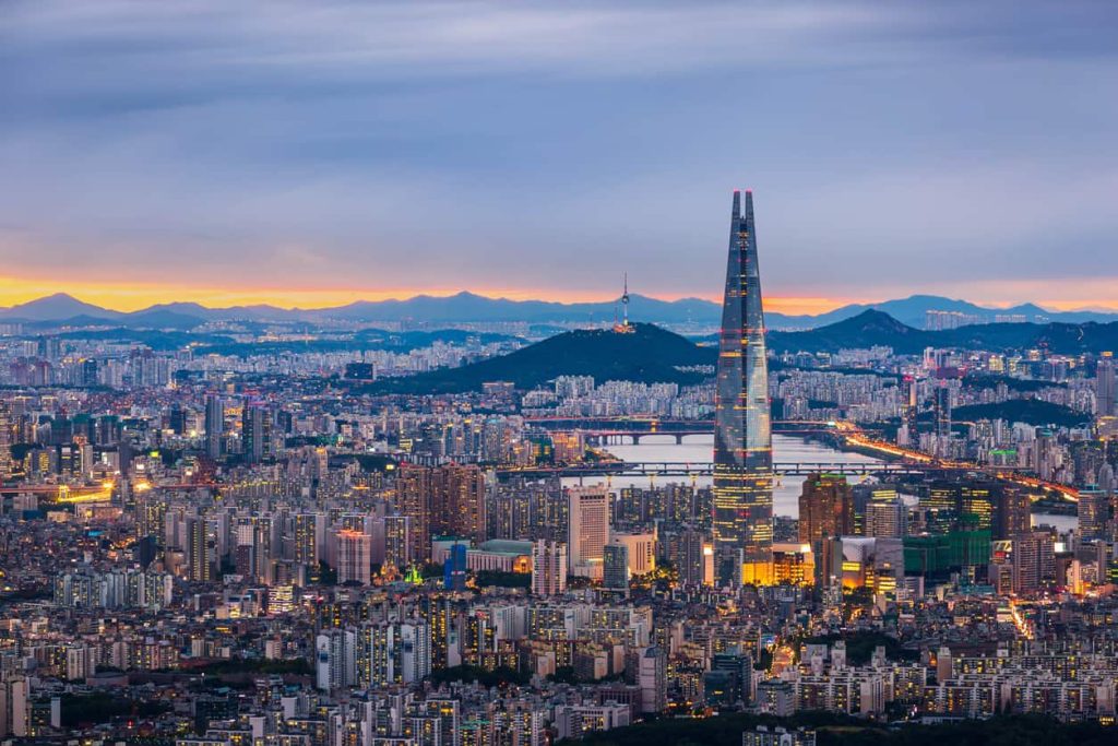 Skyline of Seoul, South Korea that has many tall buildings and lots of lights with the sun setting in the background.