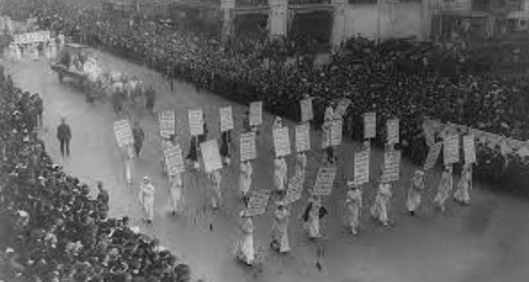 Women marching in a street with signs and with crowds on either side