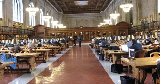 Large hall filled people reading at tables and chairs enclosed by wooden bookshelves 