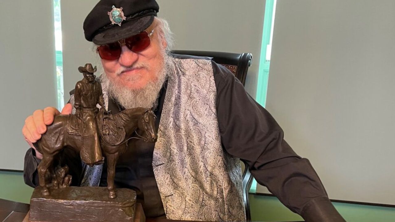 George R. R. Martin wearing sunglasses and his hand on a figurine of a man on a horse