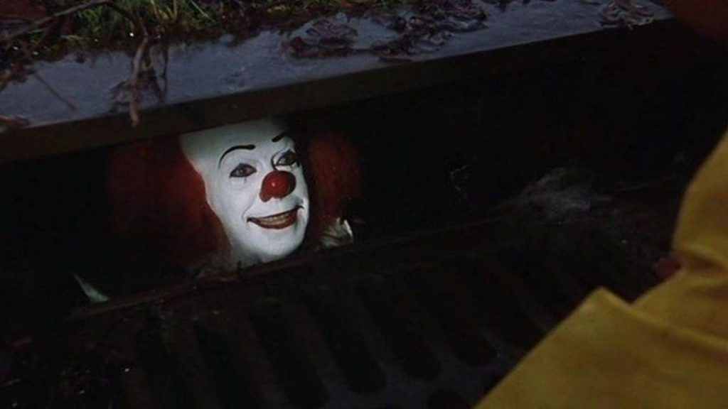 Tim Curry as Pennywise in 1990 miniseries, peeking out of a sewer drain.