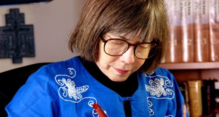 Anne Rice writing at a desk wearing a blue sweater and glasses in front of a bookshelf