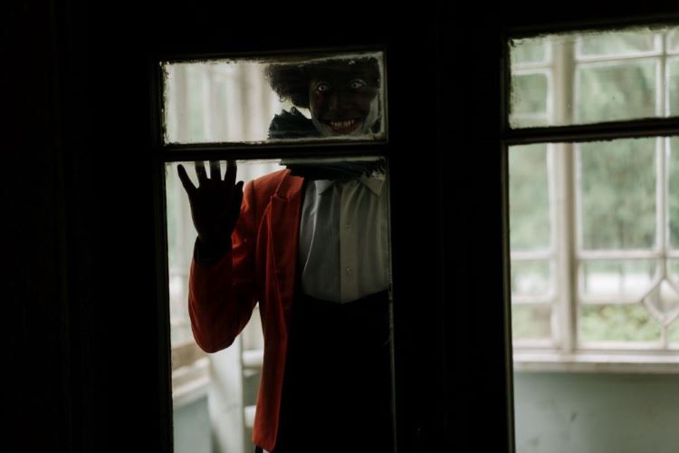 A scary figure dressed like a clown looks in through the window. A red jacket over a white shirt with a black ruff around the neck.