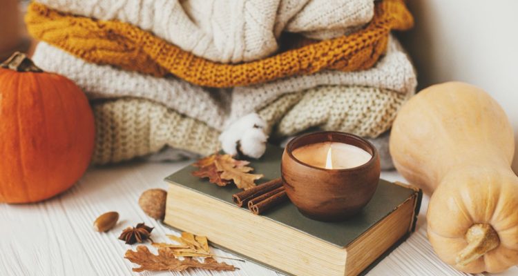 A fall themes photo that includes two pumpkins, some white and orange blankets, some leaves, a candle and a book.