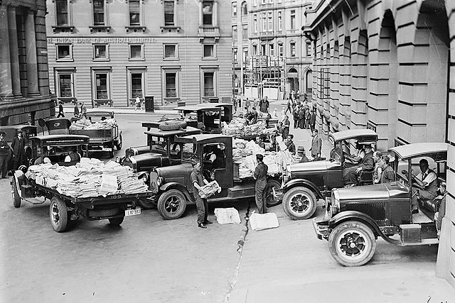 Black and white photo of newspapers being loaded into cars.