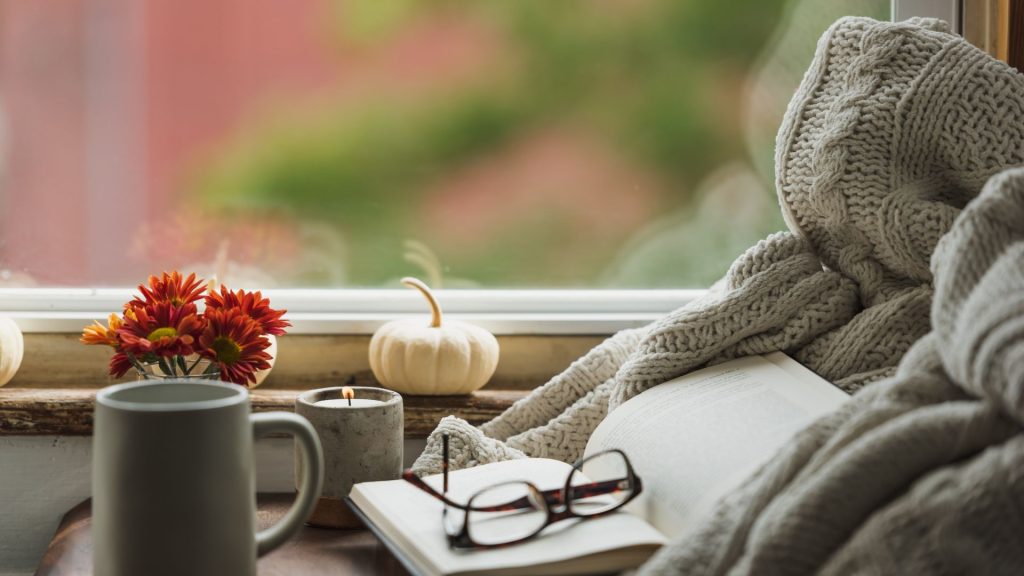 Cozy window viewing blurred autumn leaves with a blanket, glasses, coffee mug, and candle lit. 