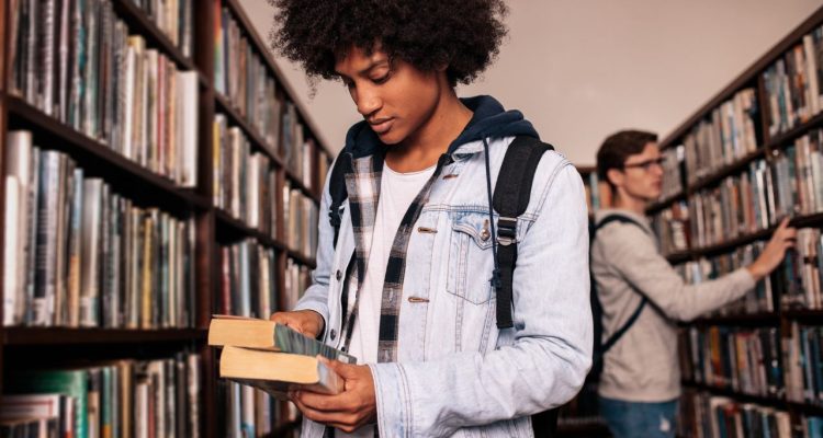 A student looking down at books in his hands, bookshelves in the background.