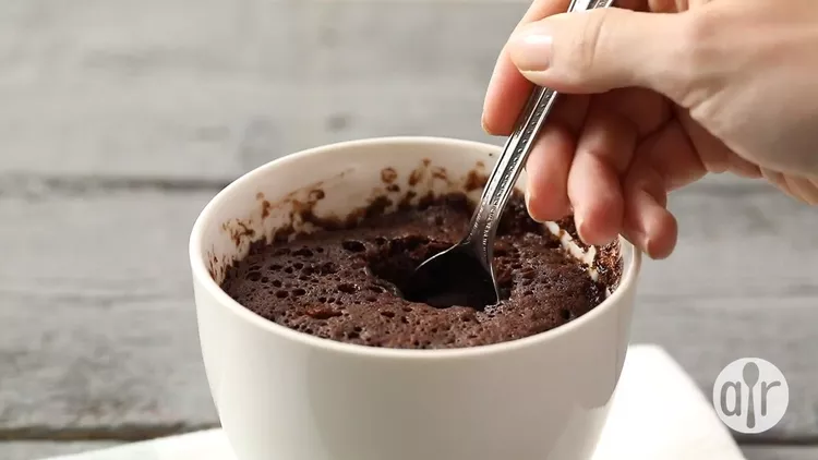 A mug cake with a hand dipping in a spoon to take a bite