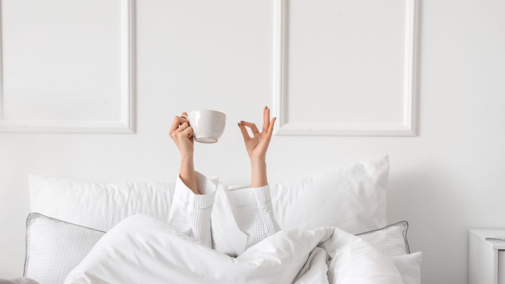 Person holding a coffee mug in the air in a cozy bed