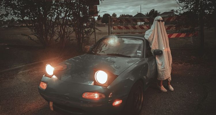 A person dressed as a ghost leaning on a car with its headlights on