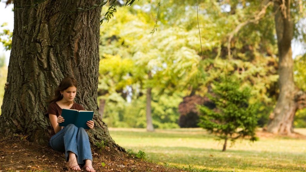 girl leaning against a tree outside reading