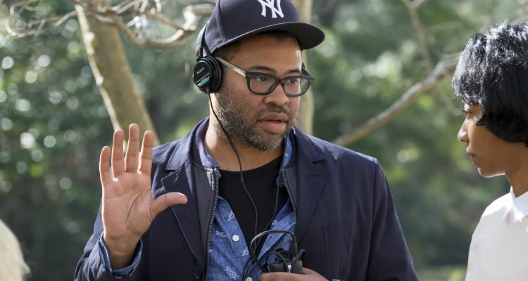 Jordan Peele directing the movie get out wearing a navy baseball cap and navy jacket and a black t-shirt in front of trees
