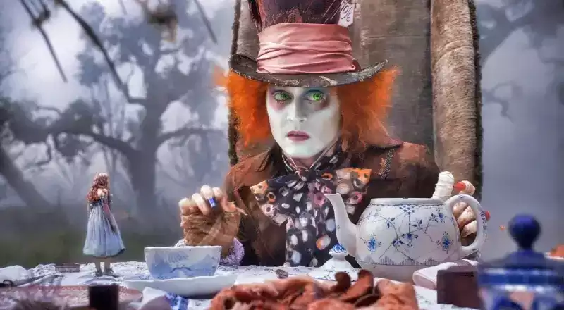 The Mad Hatter from Alice in Wonderland with a hat on as he looks at something offscreen. Alice is standing on the table in a blue dress.