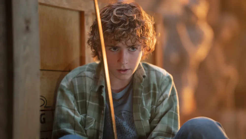 Walker Scobell as Percy Jackson wearing a grey flannel and holding a sword as he sits in front of a wooden crate.