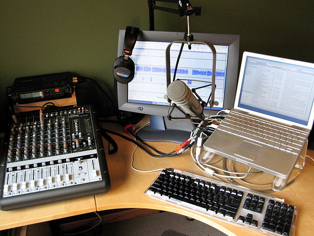 Home podcast studio with two computer moniters, a microphone, a pair of headphones, and a sound board.