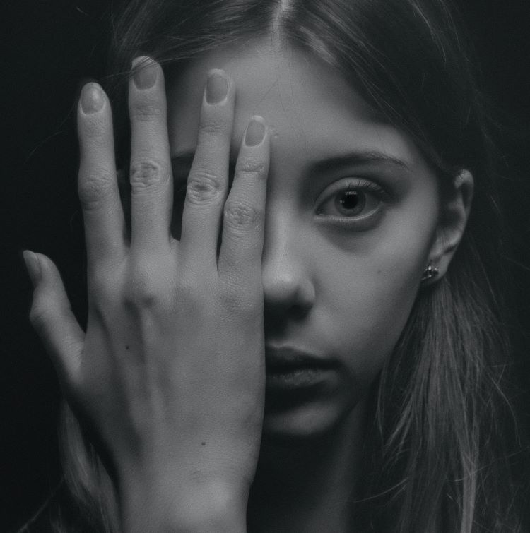 A young woman in a black and white filter. Half her face is covered by her hand. The exposed part of her face looks scared.