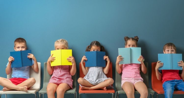 Five children sitting in a line holding up books to cover the bottom of their faces. They sit against a blue background.
