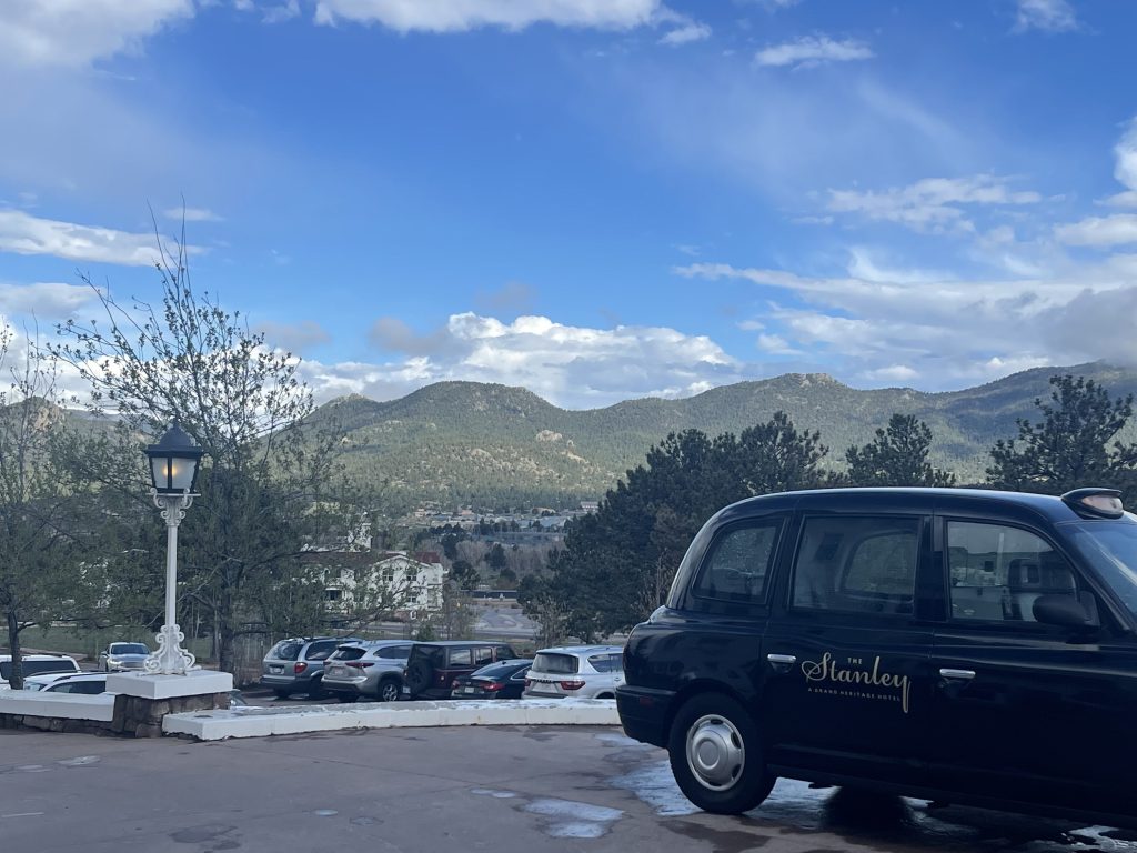 The view from the front of The Stanley Hotel. Inlcudes the mountains as well as an old school car that says "The Stanley Hotel" on it.