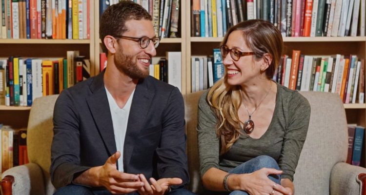 Adam Levy and Ashley Nelson Levy chatting on a couch in front of a bookshelf