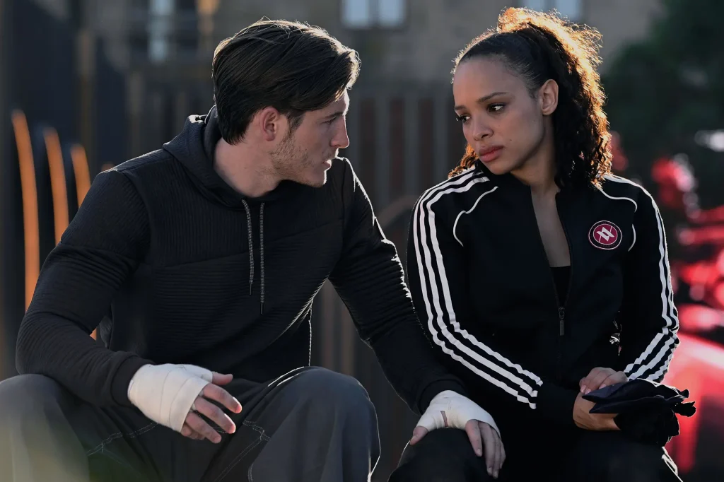Dimitri in a black sweatshirt with his hand on Rose's thigh. Rose is wearing a black training set with white strips and holding gloves in her lap.