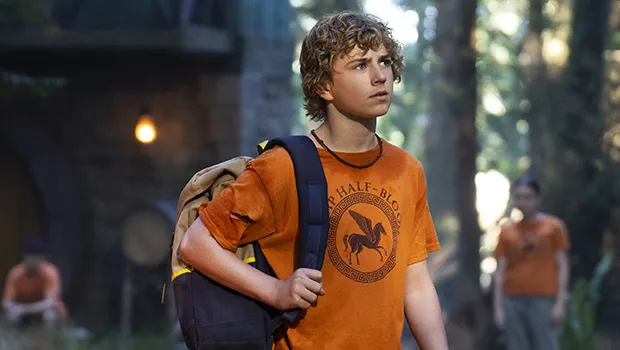 Walker Scobell as Percy Jackson with blonde hair, an orange Camp Half-blood shirt, and a black and tan backpack over his shoulder.