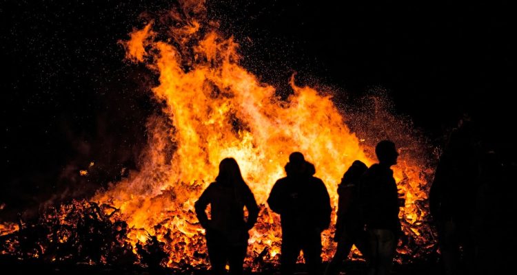 A large bonfire blazes at night while a group of people gather around it