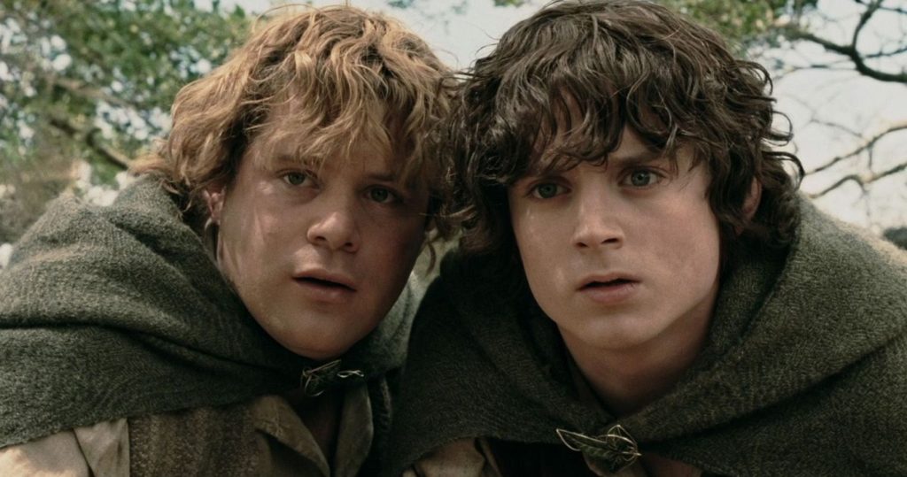 Sam is on the left and Frodo is in on the right. They are standing close together and looking at something or spying on something or someone. They are outside. There are branches behind their heads. They look with cautious faces.