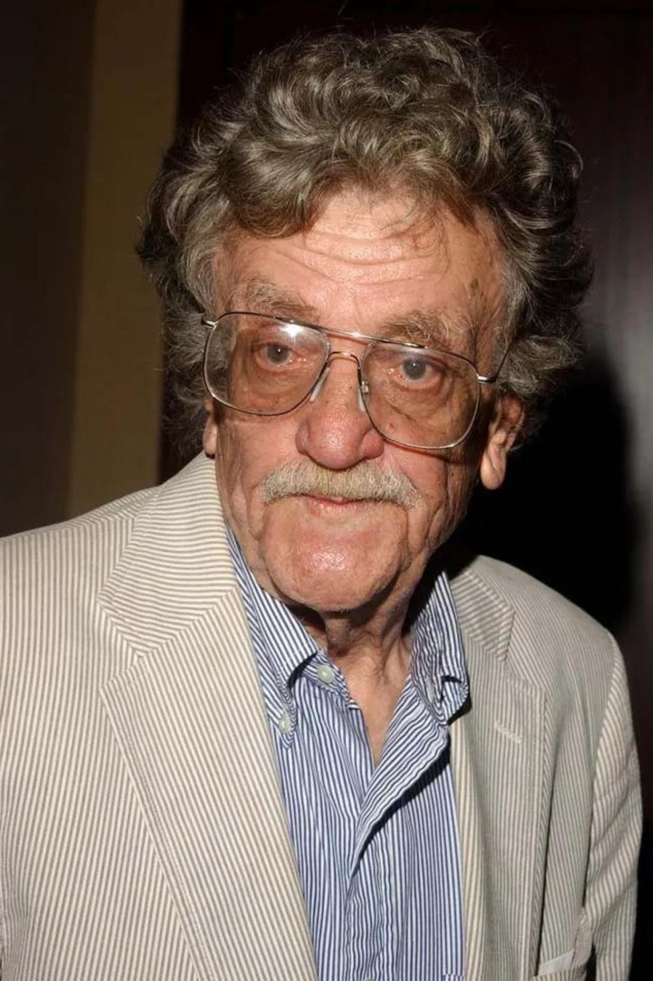 An older Kurt Vonnegut is wearing a beige striped suit jacket with a gray-blue striped buttoned shirt. He stands in front of a brown background.
