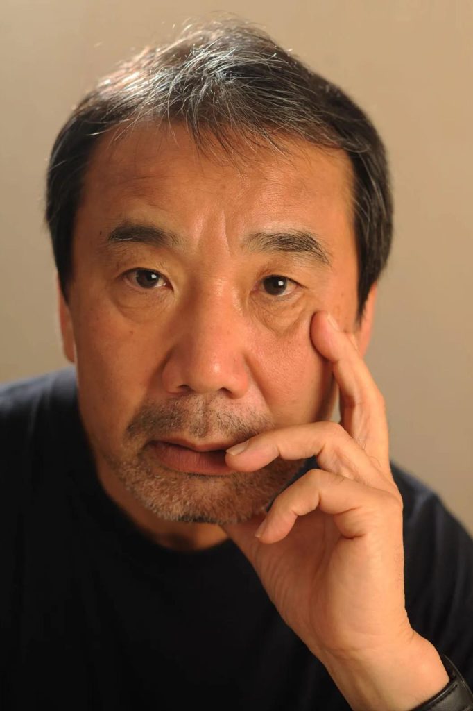 Haruki Murakami is dressed in a black top. His head sits in his hand, and he looks pensive. He is in front of a brownish-beige background.