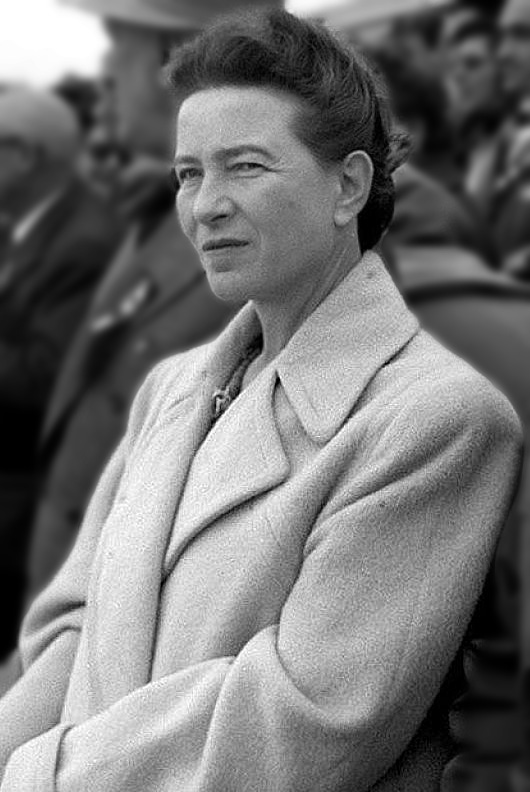 Black and white of Simone de Beauvoir. She is wearing a white coat with her hair pulled up. There's a blur of people behind her.