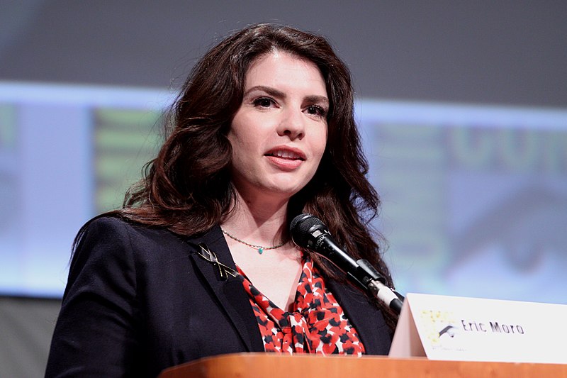 Color photo of Stephenie Meyer. She's wearing a blue blazer with a patterned red blouse. There's a microphone in front of her. Her hair is down around her shoulders.