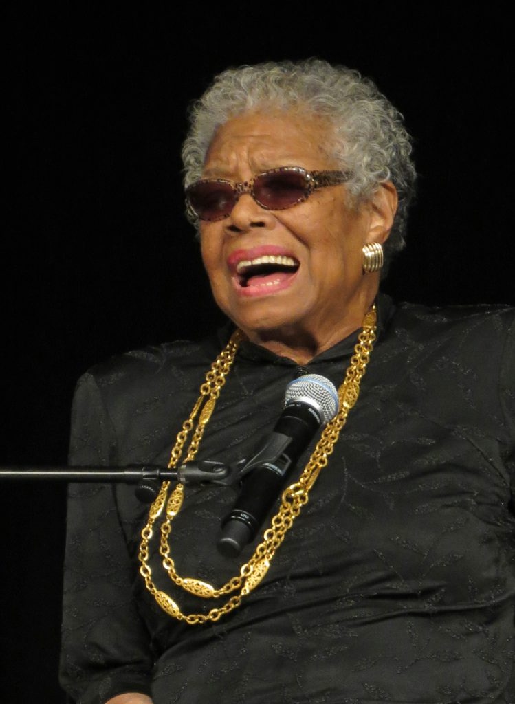 An older Maya Angelou is wearing a black top with a long, gold necklace. She also wears dark glasses. A microphone is set up in front of her.