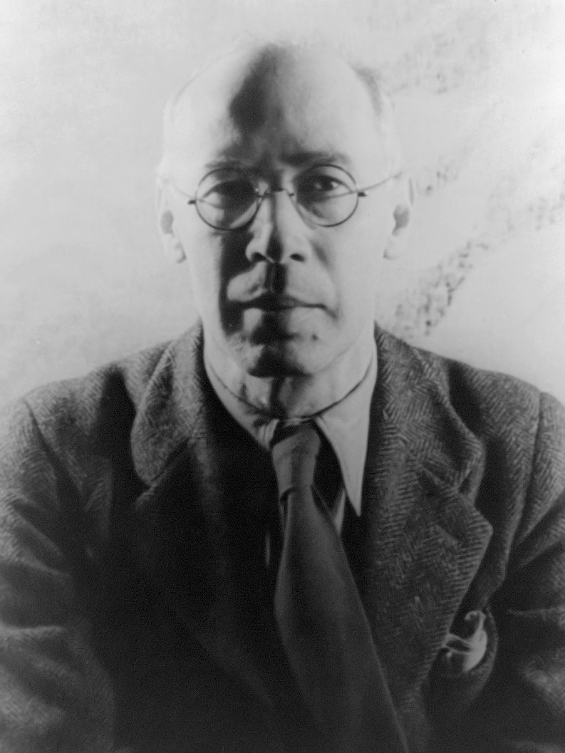 Black and white of Henry Miller. He is wearing a suit and tie. He is sitting in front of a white background.