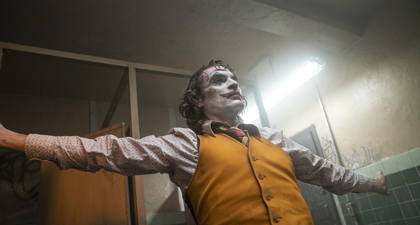 The Joker stands in a bathroom with his arms extended. The bathroom behind him is in poor condition to reflect his mental state.