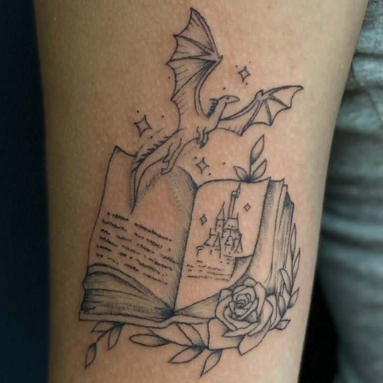 tattoo of a book with words, a castle, a dragon and a rose on an arm