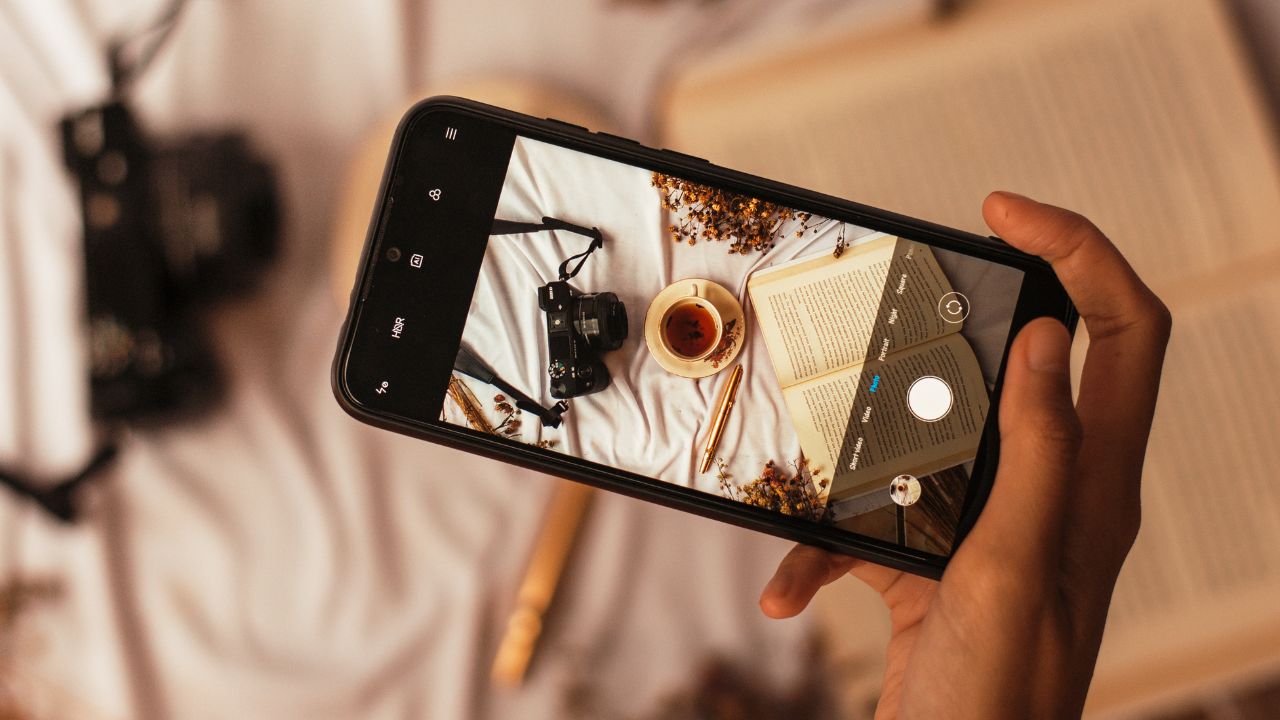 Black mobile phone takes sideways photo of a white bedspread with a camera and book laying on it.